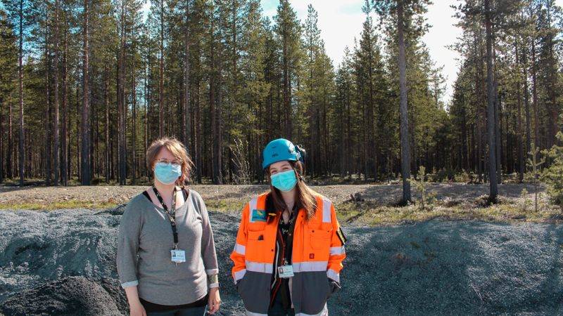 Kesäharjoittelijat poseeraavat metsätaustan edessä. Vasemmalla Lilja Ojala, joka on pukeutunut toimistovaatteisiin. Oikealla Nella Hietanen, jolla on yllään kirkkaan oranssi työtakki ja sininen kypärä.